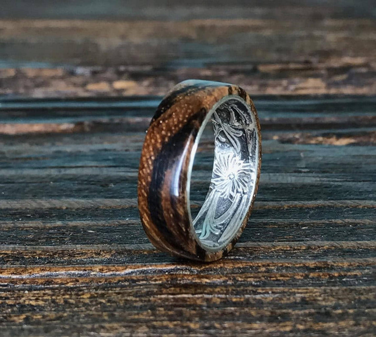 A zebrawood ring with a silver interior and engraved floral pattern is shown from a slightly different angle on a dark wooden surface, highlighting the wood's stripes.