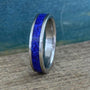 A silver-colored titanium ring with a central inlay of crushed blue lapis lazuli, viewed from a slight angle on a wooden surface.