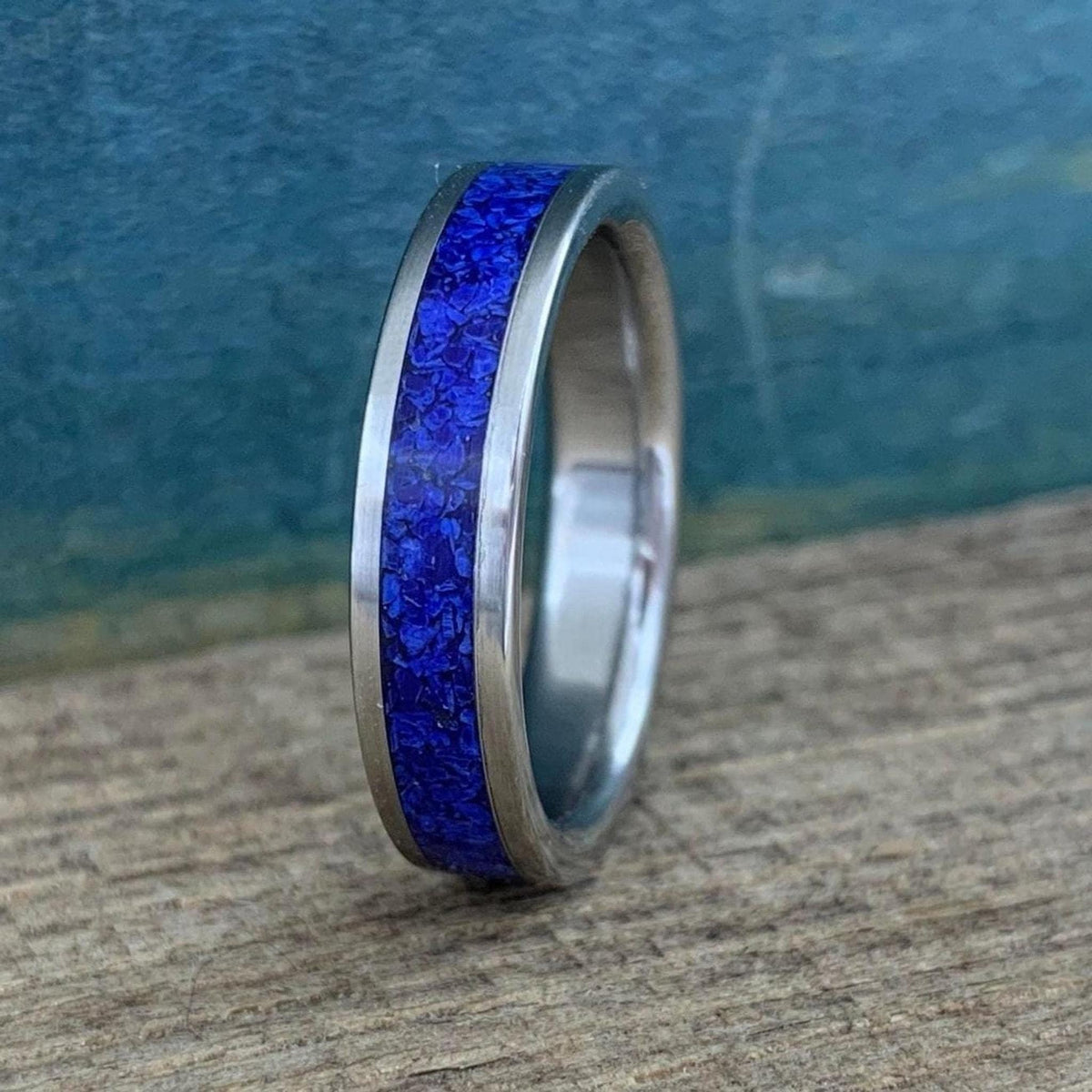 A silver-colored titanium ring with a central inlay of crushed blue lapis lazuli, viewed from a slight angle on a wooden surface.