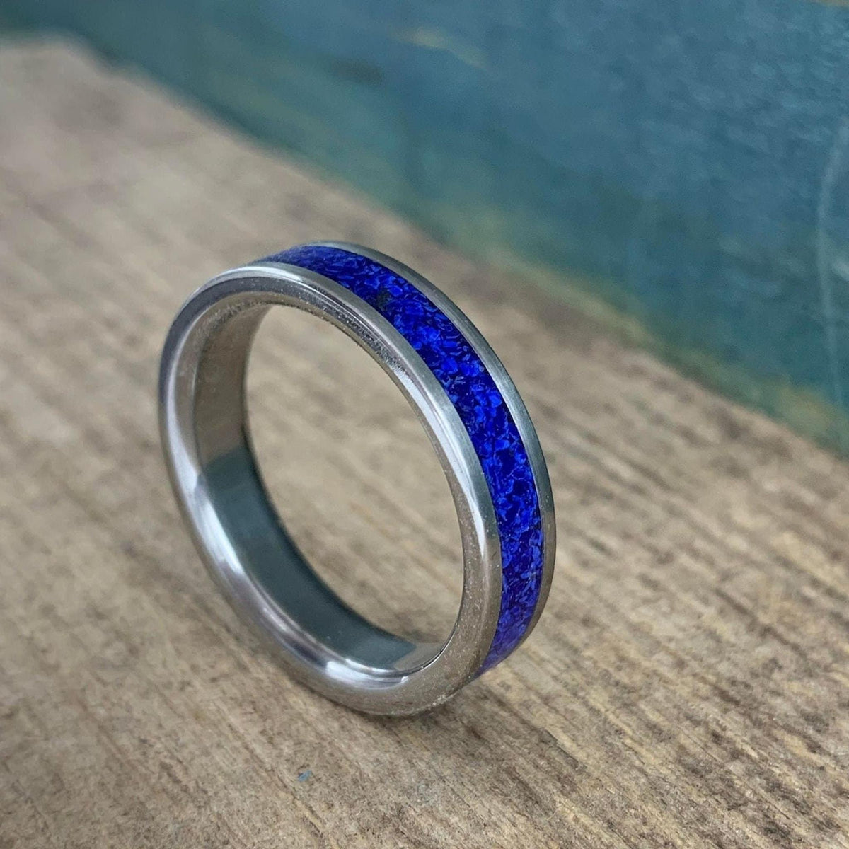 A silver-colored titanium ring with a central band of crushed blue lapis lazuli, shown at an angle on a light wooden surface.
