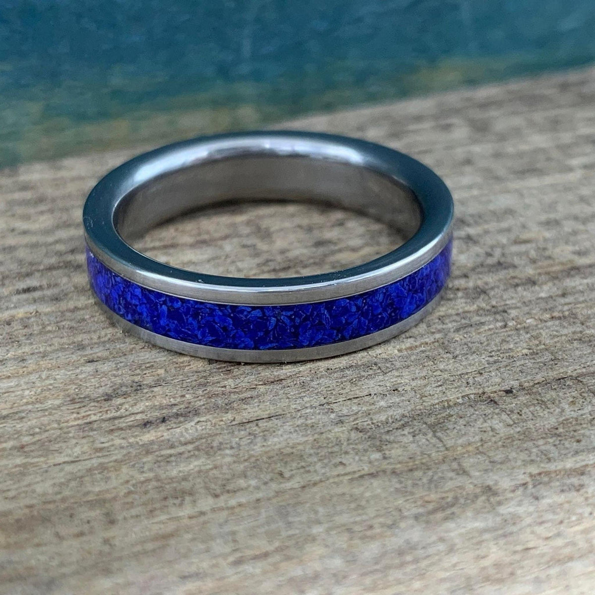 A silver-colored titanium ring with a central inlay of crushed blue lapis lazuli, viewed from a slight angle on a light wooden surface.