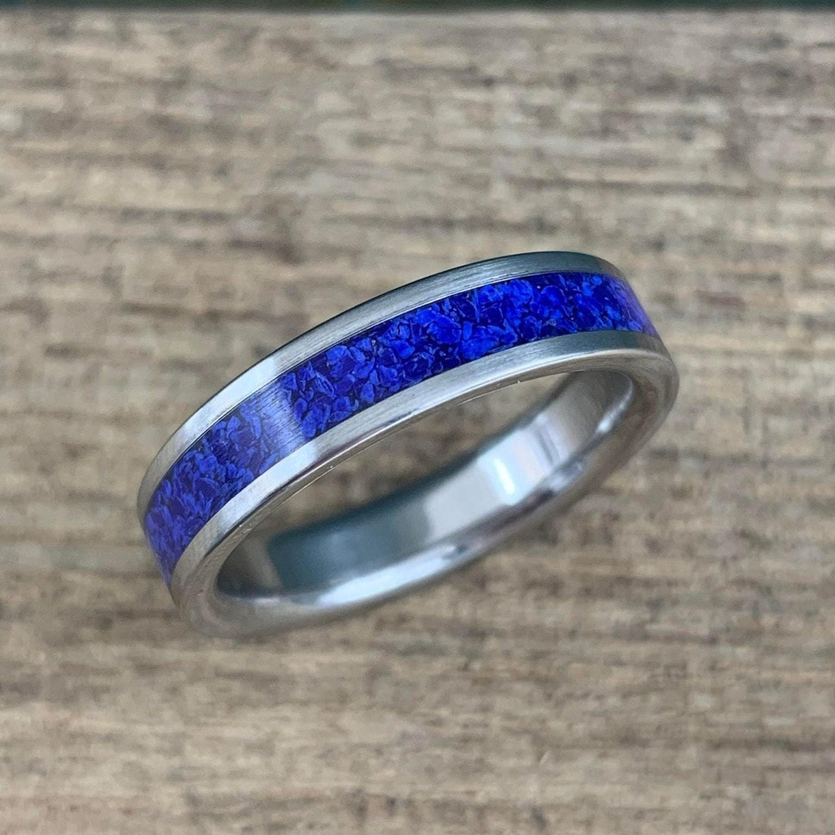 A silver-colored titanium ring with a central inlay of crushed blue lapis lazuli, viewed from a slight angle on a light wooden surface.
