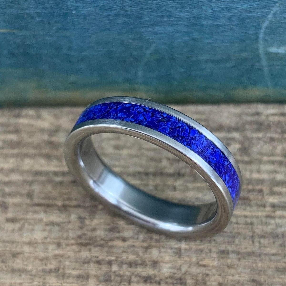 A silver-colored titanium ring with a central band of crushed blue lapis lazuli, viewed from a slight angle on a light wooden surface.