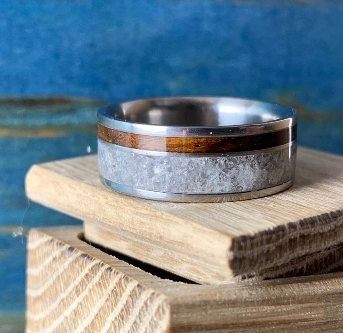 A titanium ring with a central band of white rose quartz and a narrow strip of dark kingwood, resting on a light wooden block.