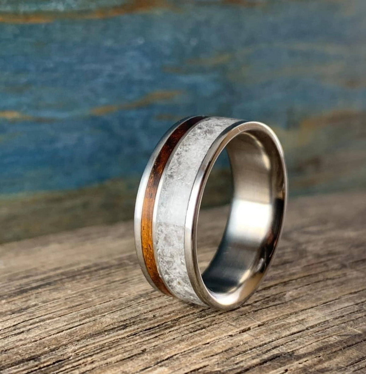 A titanium ring with a central band of white rose quartz and a narrow strip of dark kingwood, shown at an angle on a wooden surface.