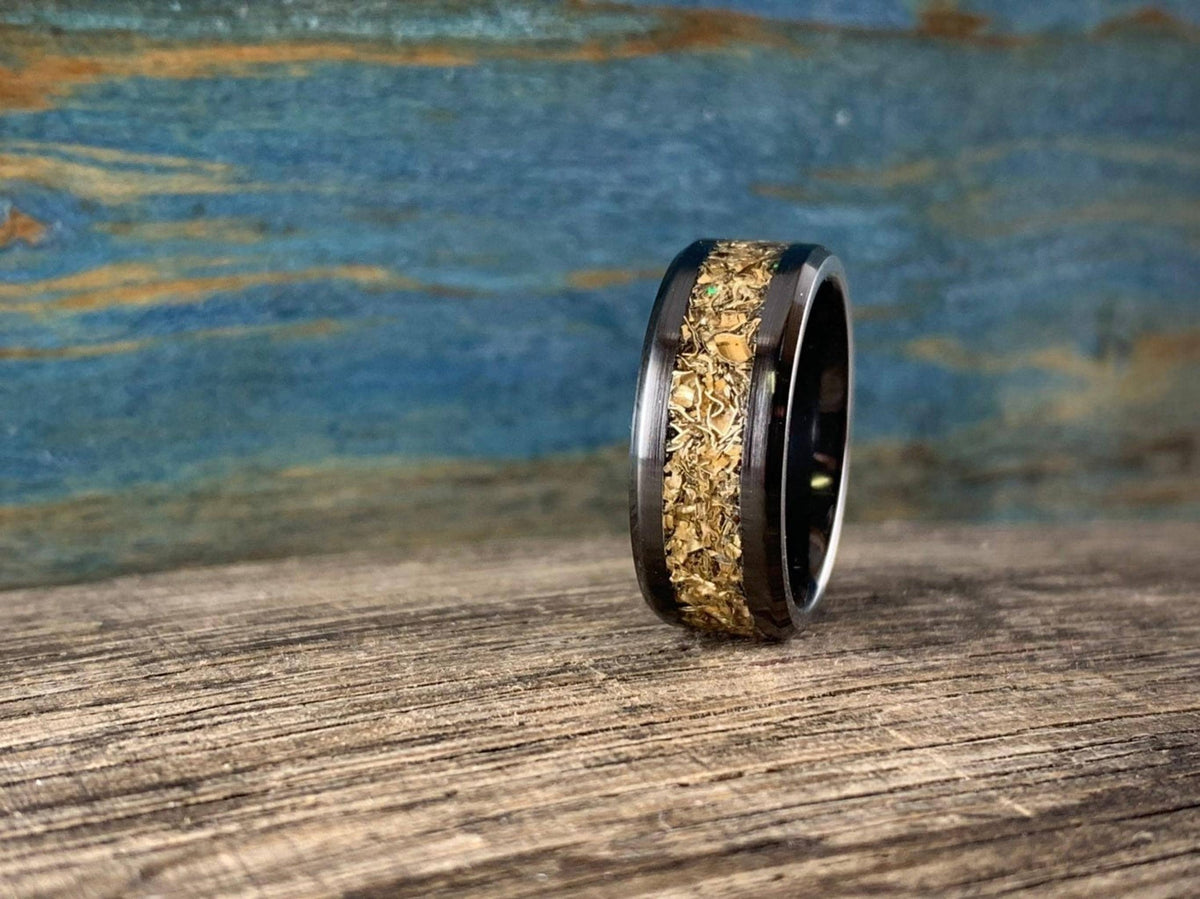 A black ceramic ring with a textured gold-colored inlay is shown from the side on a wooden surface with a blue background.