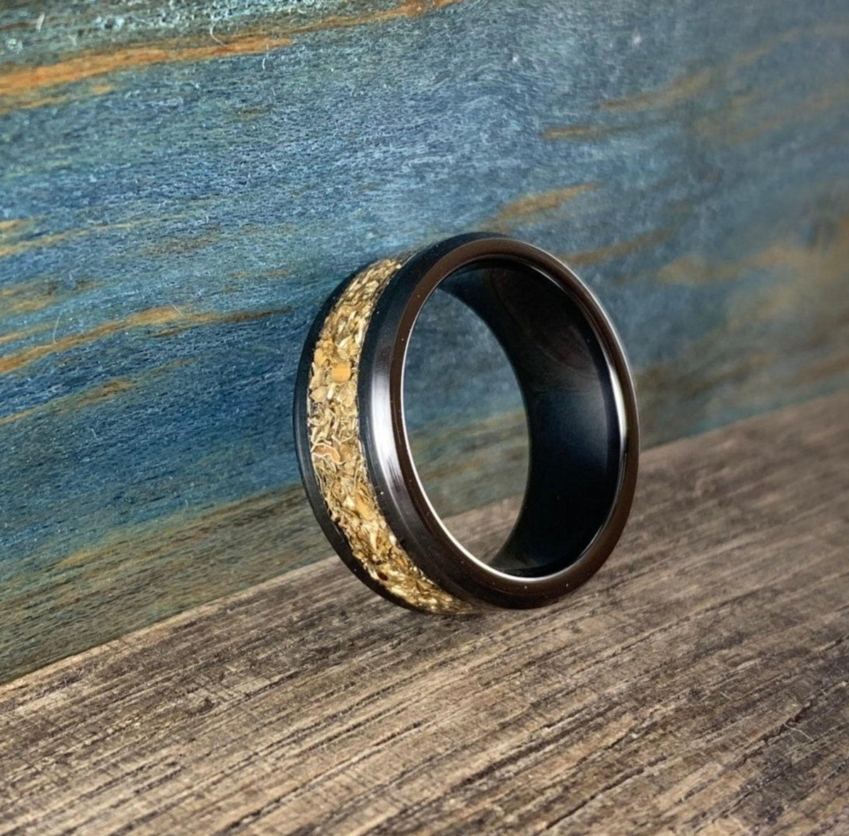 A black ceramic ring with a textured gold-colored inlay is shown from the side on a wooden surface with a blue background.
