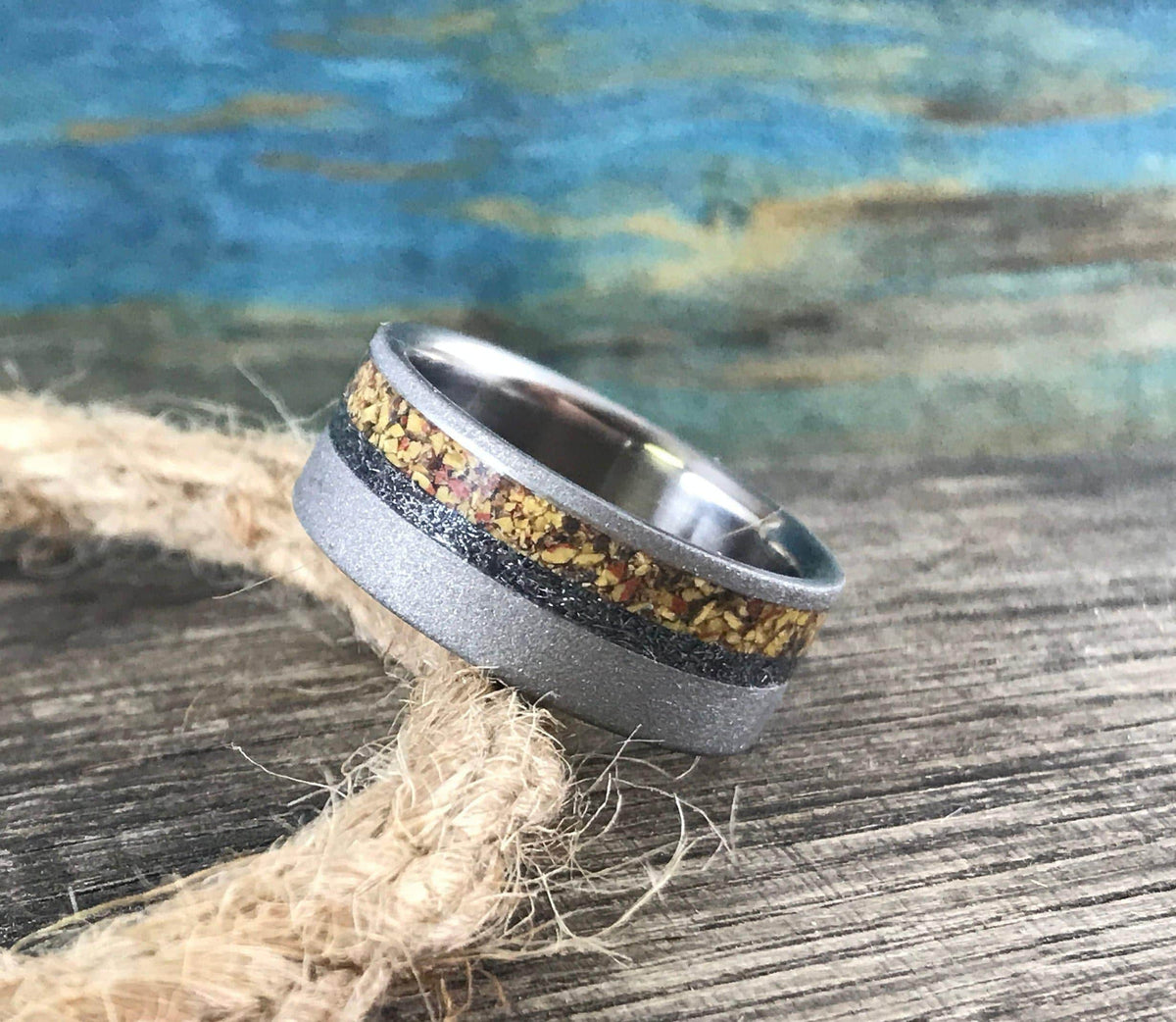 A silver-colored ring with a sandblasted finish and a central band of yellow, orange, and black flecks, rests on a coiled rope.