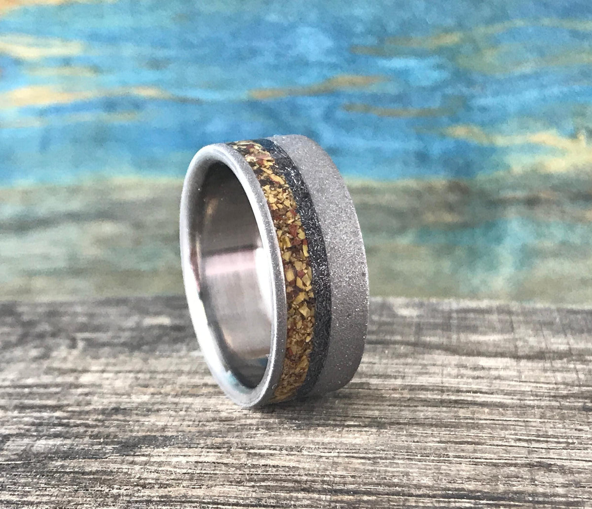A silver-colored ring with a sandblasted finish and a central band of yellow, orange, and black flecks, is viewed from a slight angle on a wooden surface.