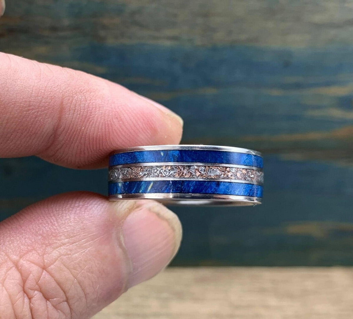 A hand holds the metal ring with blue wood and metallic inlays against a rustic background.