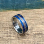 A metal ring featuring a blue wood and metallic textured inlay sits angled on a wood surface.