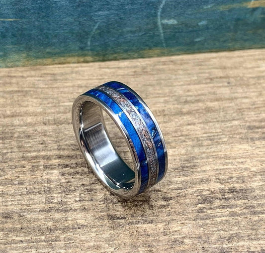 A metal ring featuring a blue wood and metallic textured inlay sits angled on a wood surface.