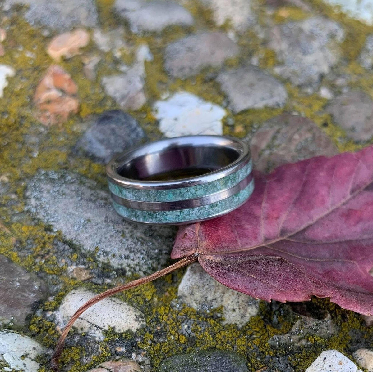 A moss agate and titanium ring lies beside a purple leaf on a bed of grey stones covered in moss.