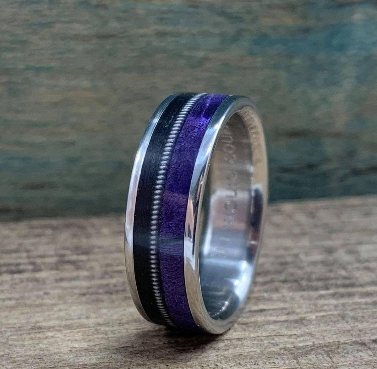 A close-up shot of the titanium ring showcasing its textured guitar string and purple wood inlay against a rustic wooden background.