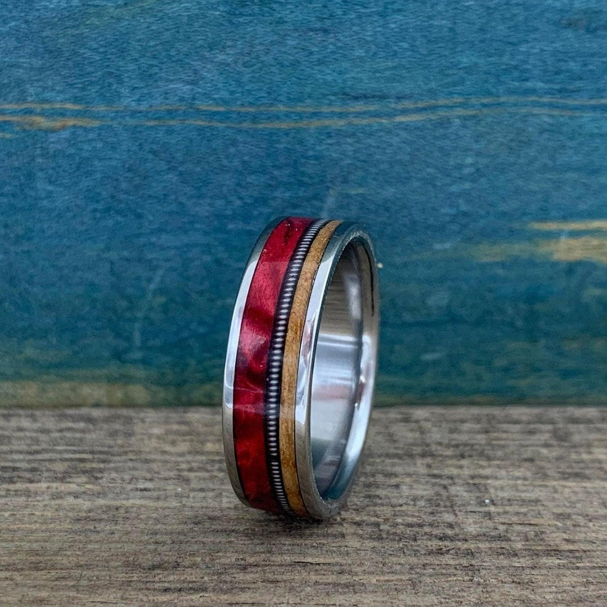 A custom ring rests upright on a rustic wooden surface against a blue textured backdrop, showing silver metal edges framing red wood, tan wood, and a coiled guitar string inlay.