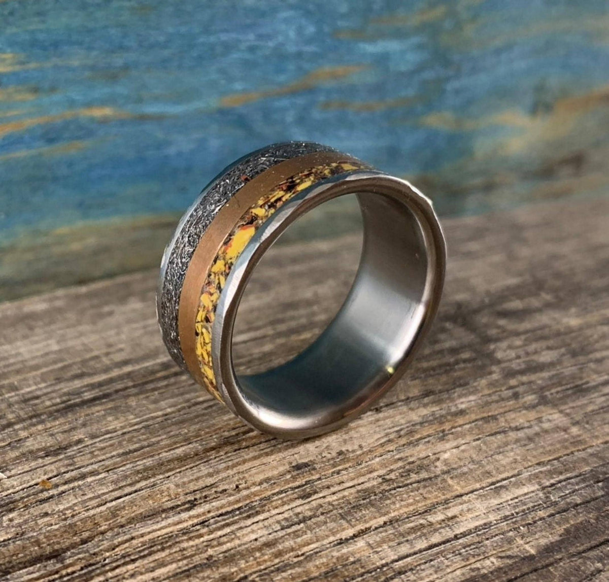 A silver-colored ring with a hammered texture on the outer edges, a smooth copper-colored band, and a textured yellow-orange band in the center, viewed from a low angle.