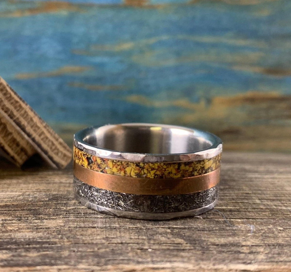 A silver-colored ring with hammered outer edges, a smooth copper-colored band, and a textured yellow-orange band in the center, with a wooden object in the background.