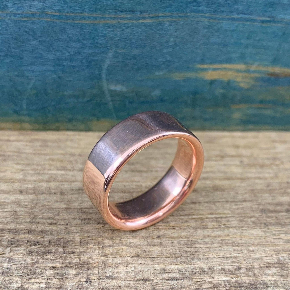 A matte rose gold-colored ring with a slightly rounded top surface is angled on a wooden surface.