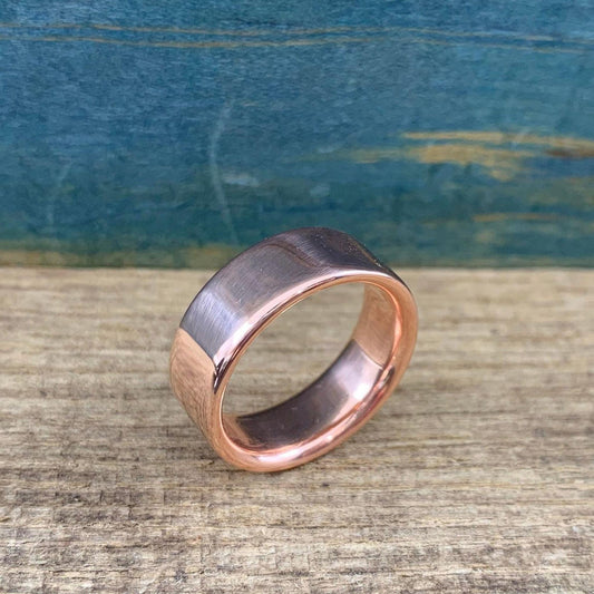 A matte rose gold-colored ring with a slightly rounded top surface is angled on a wooden surface.