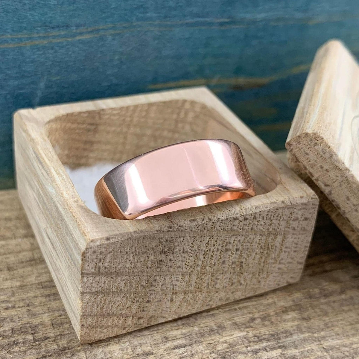 A shiny rose gold-colored ring with a flat top surface rests inside an open light wood box.