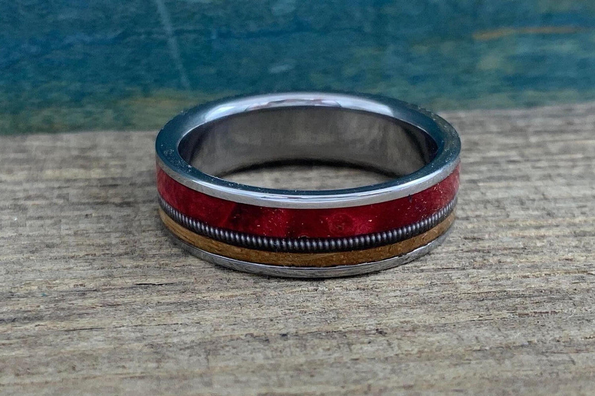 The ring is positioned horizontally with the silver edges framing the center inlays of red wood, coiled string, and tan wood against the rustic background.