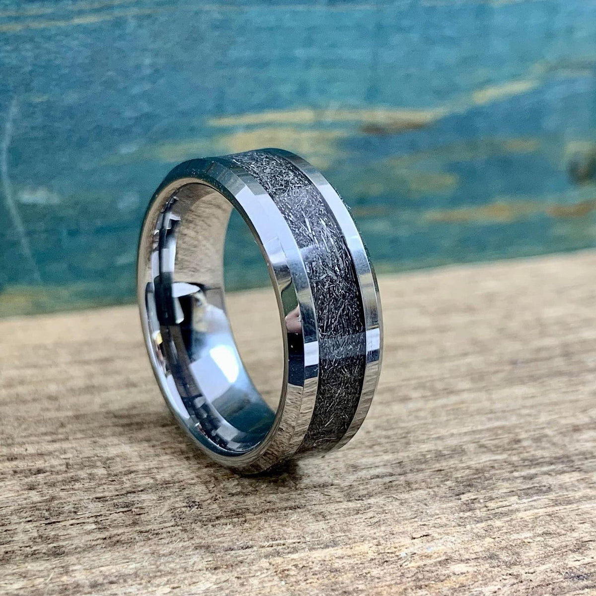 A silver-toned tungsten ring with a dark, textured meteorite inlay is centered on a wooden surface, showing the top and side.