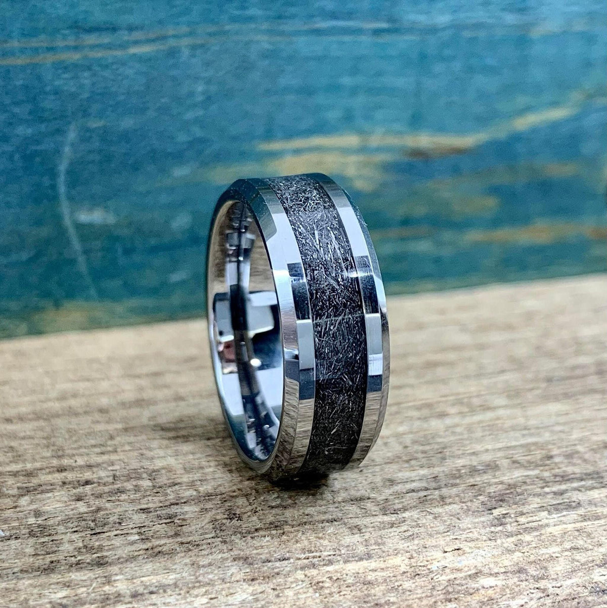A silver-toned tungsten ring with a dark, textured meteorite inlay is shown from a slightly elevated angle on a wooden surface.