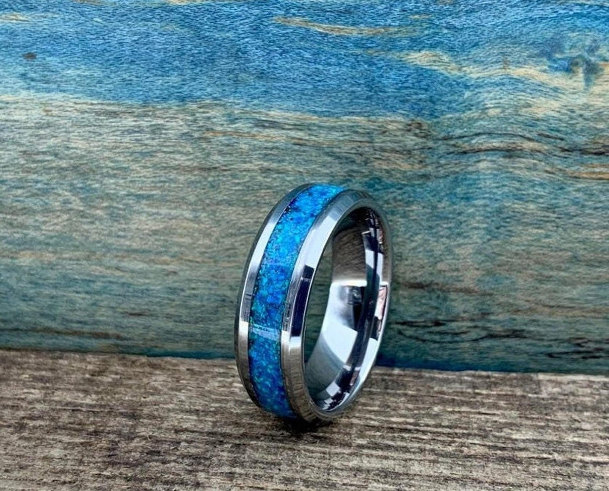 A silver-colored ring with a blue-green opal inlay is shown from a slightly different angle on a wooden surface.