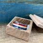 A silver-colored ring with red, white, and blue stripes is displayed inside an open, light-colored wooden box.