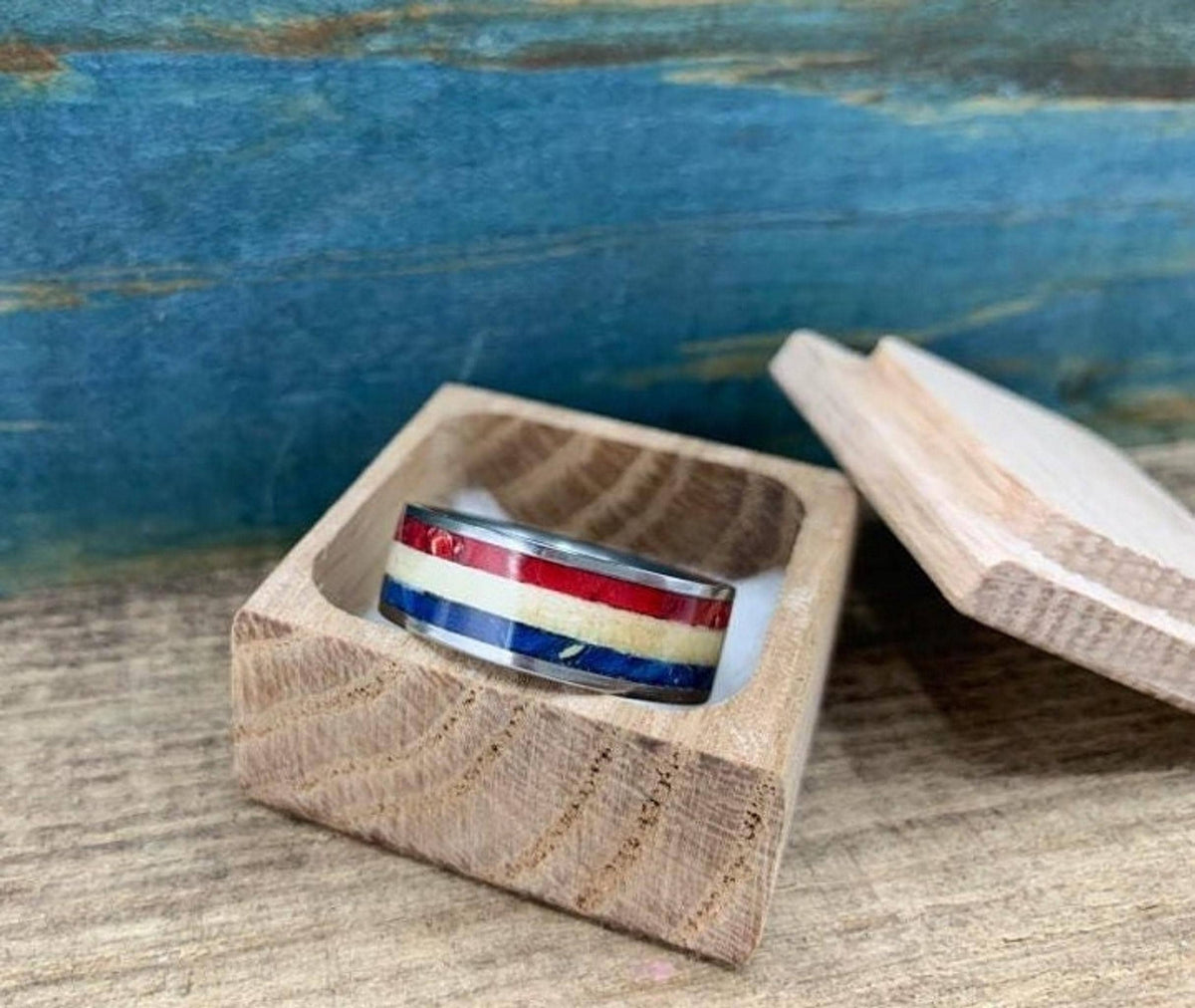 A silver-colored ring with red, white, and blue stripes is displayed inside an open, light-colored wooden box.