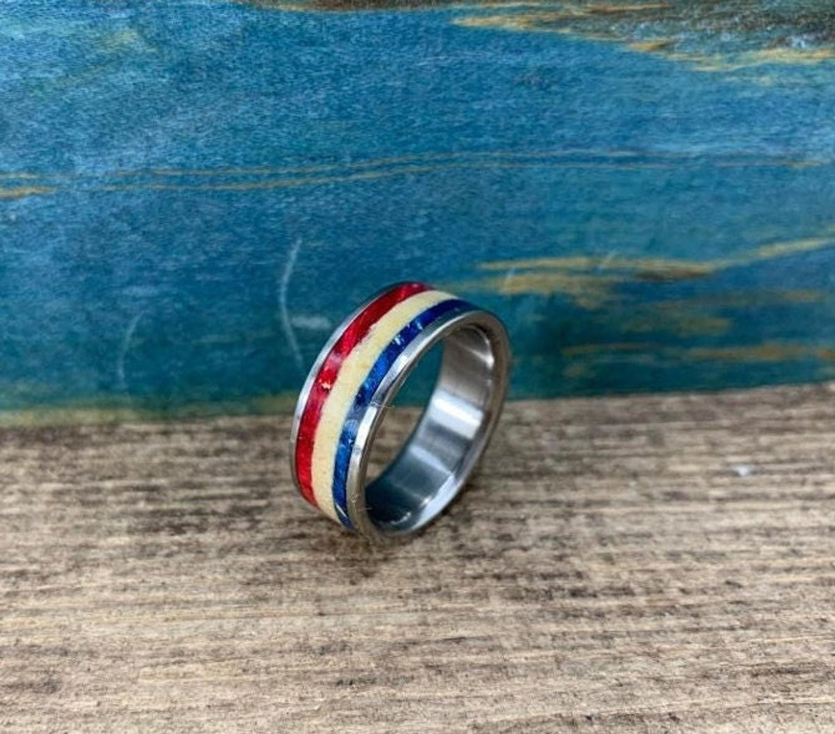 A silver-colored ring with red, white, and blue stripes is shown from a slightly elevated angle, slightly to the right, on a wooden surface.
