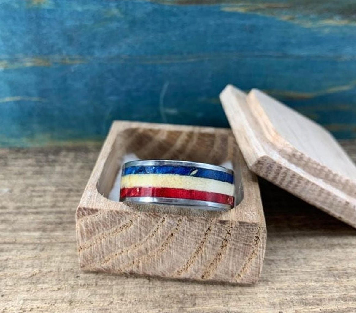A silver-colored ring with red, white, and blue stripes is displayed inside an open, light-colored wooden box, with the lid next to it.