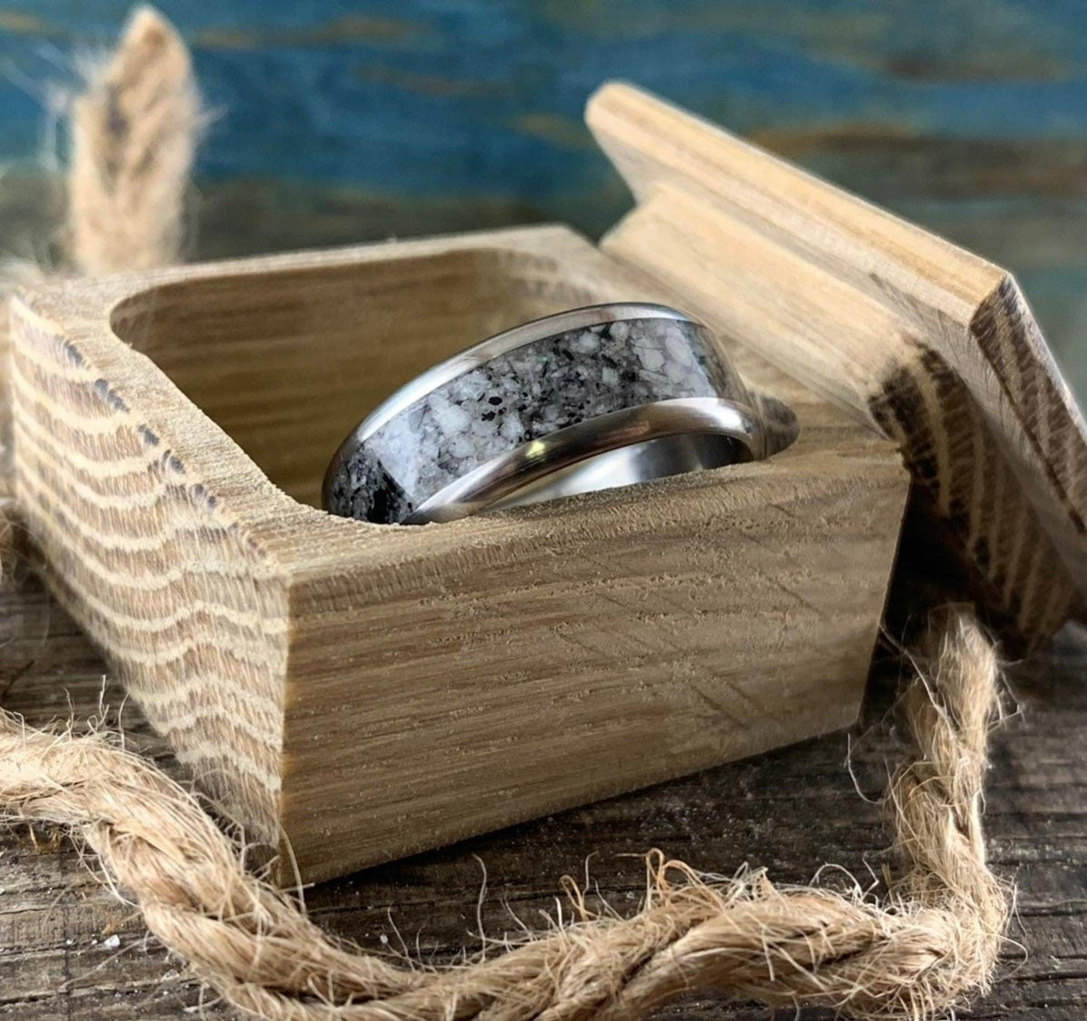 A silver-colored ring with a speckled gray and white granite inlay is nestled inside an open wooden box, with twine in the foreground.