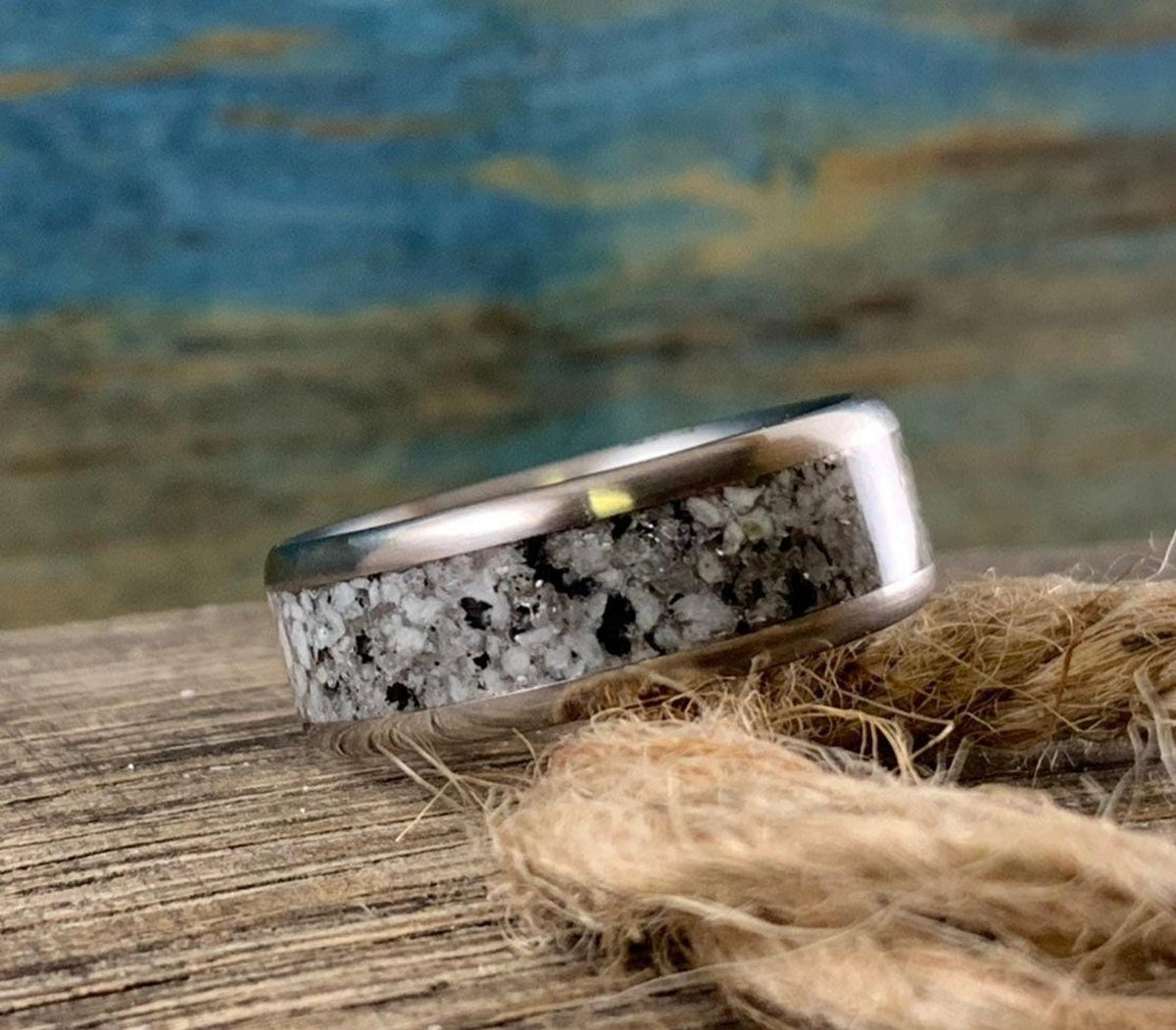 A silver-colored ring with a speckled gray and white granite inlay is positioned on a coil of natural twine.