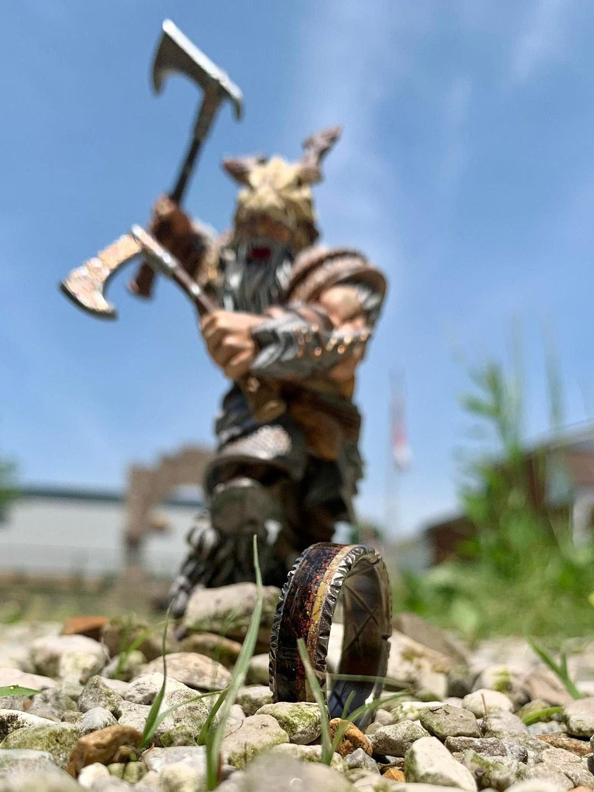 A textured wedding band rests on gravel outdoors with a blurred Viking action figure holding axes in the background.