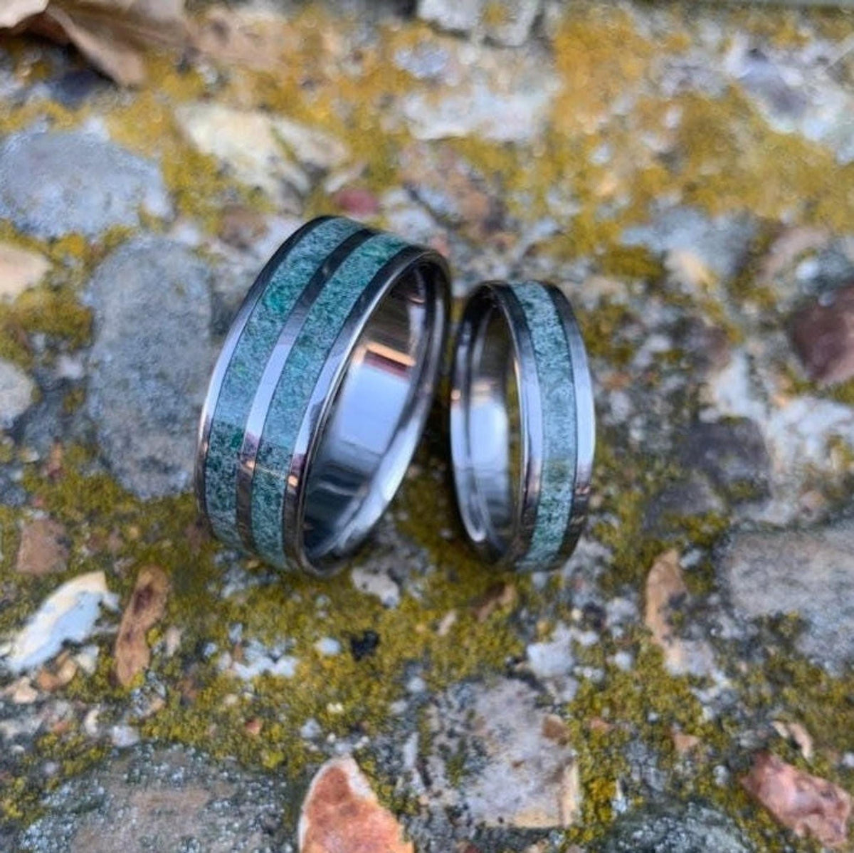 Two metal rings with green moss agate inlays rest upright side by side on a textured stone surface covered in moss.