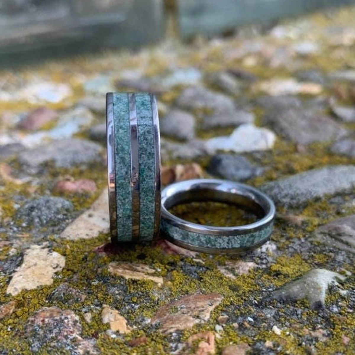 Two metal bands with green stone inlay stand upright on a mottled stone and moss surface.