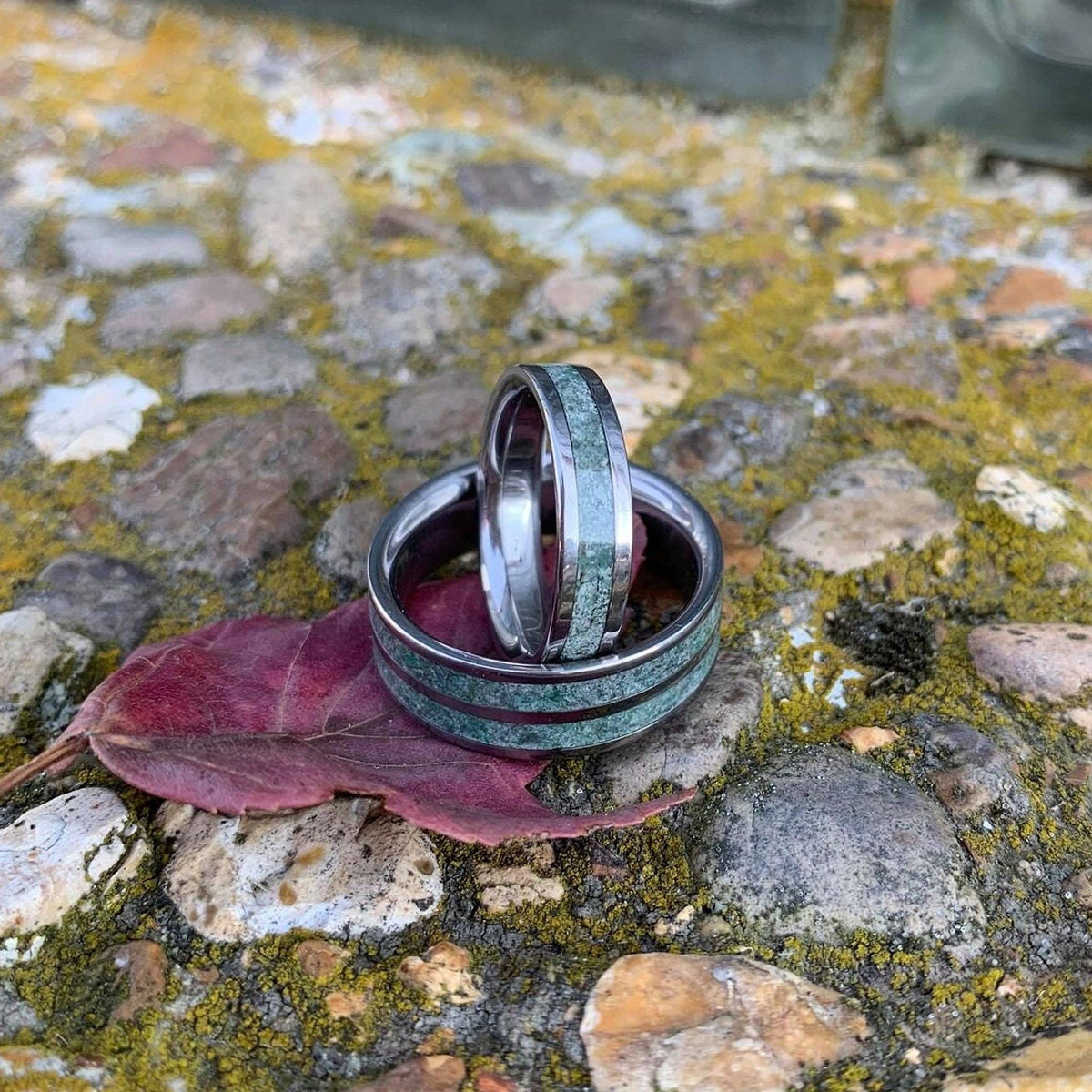 A wide moss agate ring and a narrower one rest on a dry reddish leaf against a moss-covered rock backdrop.