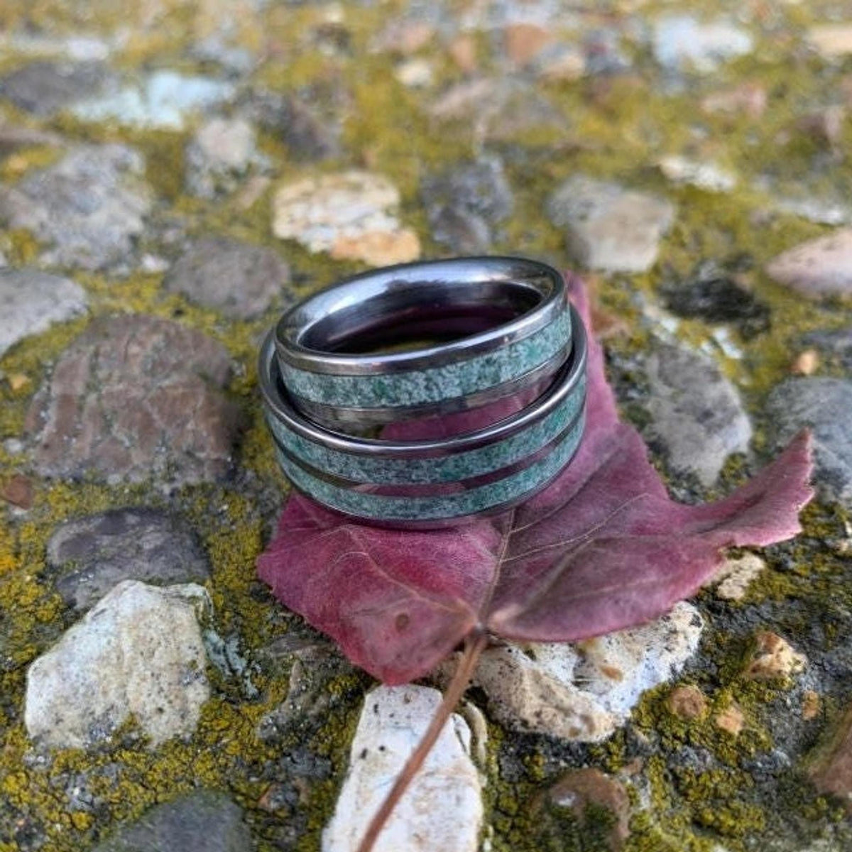 Two moss agate inlaid rings rest on a single maroon leaf over textured stones with yellow moss.