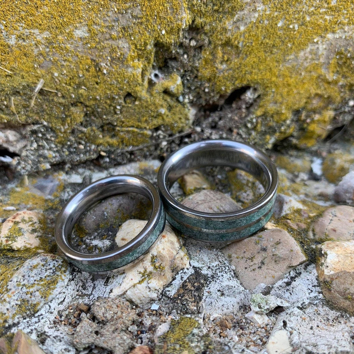 Two metal wedding bands featuring green moss agate inlays are positioned on a rocky, mossy surface.