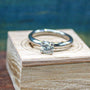 A silver-colored ring with a prong-set round gemstone sits upright on a light-colored wooden block against a blurred blue background.