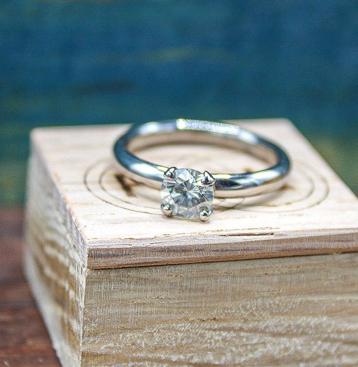 A silver-colored ring with a prong-set round gemstone sits upright on a light-colored wooden block against a blurred blue background.