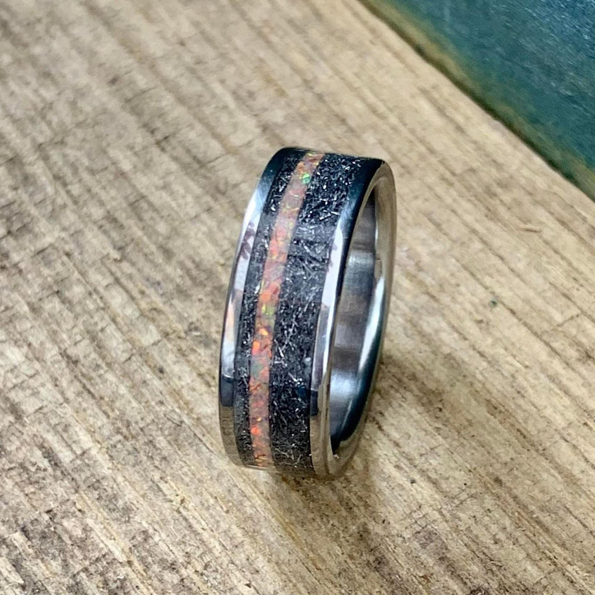 Upright view of the titanium ring showcasing its polished silver edges, meteorite inlay, and central orange opal stripe on a wooden background.