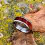 A titanium ring featuring a polished wood inlay and hammered edges, resting on mossy stones beside a dry leaf.