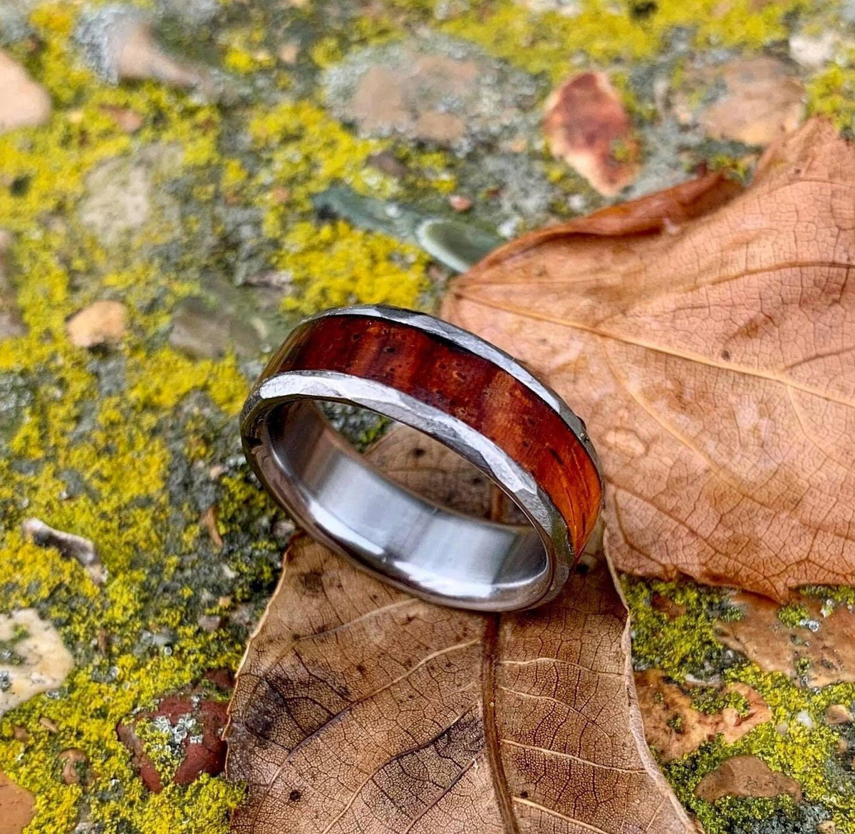 A titanium ring featuring a polished wood inlay and hammered edges, resting on mossy stones beside a dry leaf.