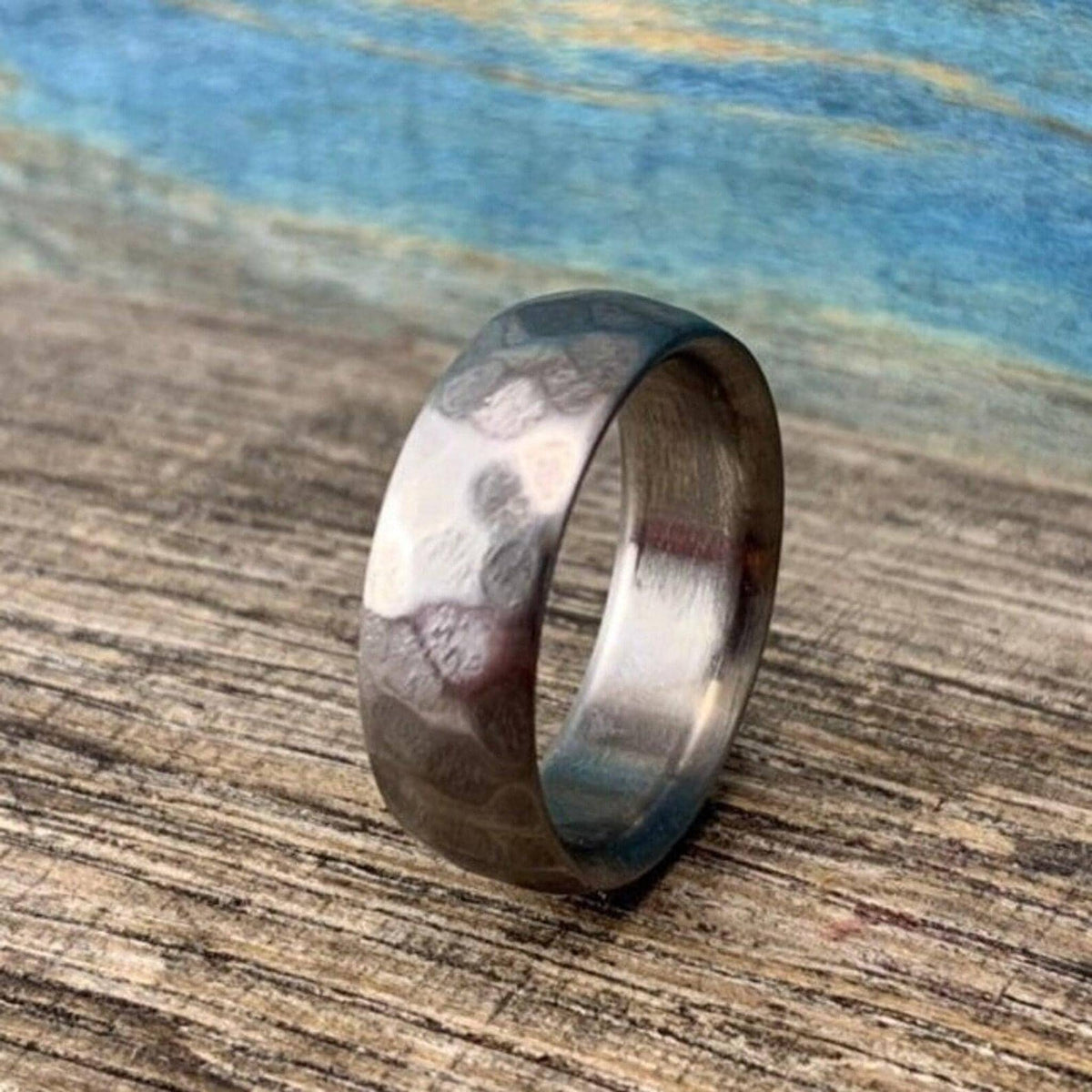 A silver-colored hammered ring angled slightly upward on a rustic wooden board with a mottled blue background.