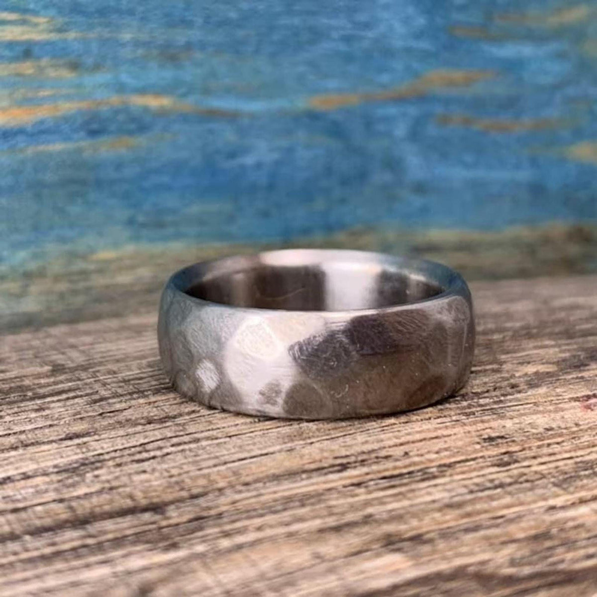A silver-colored hammered titanium band lying flat on a textured wooden surface against a blue backdrop.