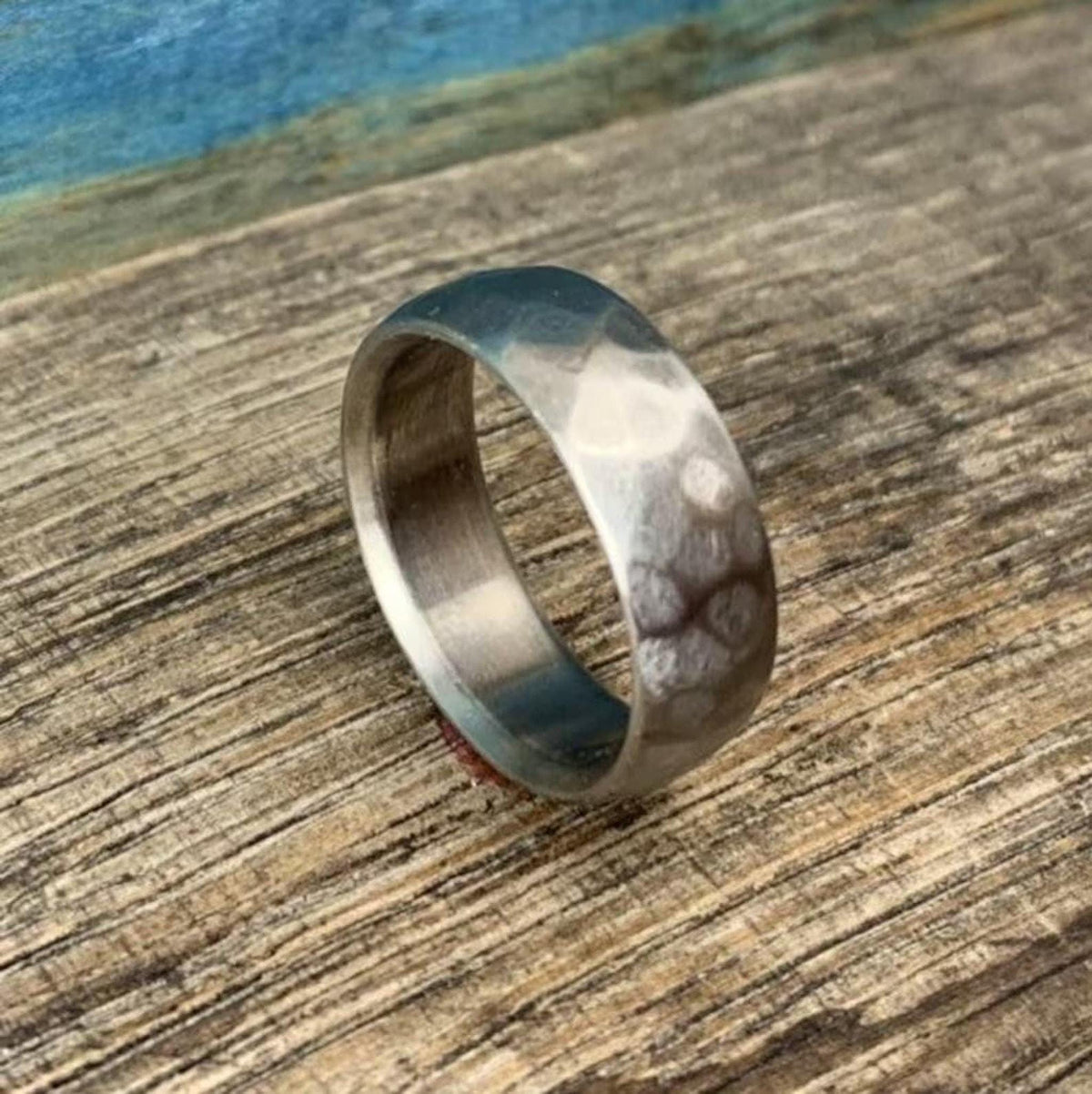 A silver-colored hammered titanium ring angled on a rustic wooden surface with a blue background.