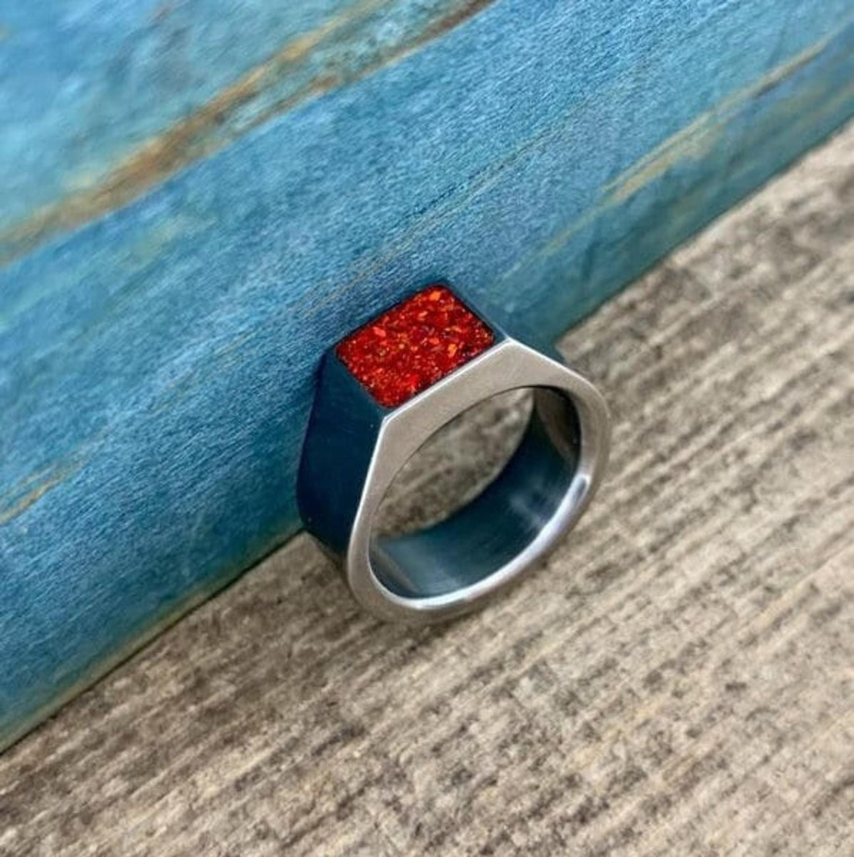 A silver metal ring with a bright orange opal square face leans against a textured blue and wood background.