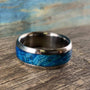 A metal ring with a wide blue wood burl inlay resting diagonally on a rustic wooden surface.